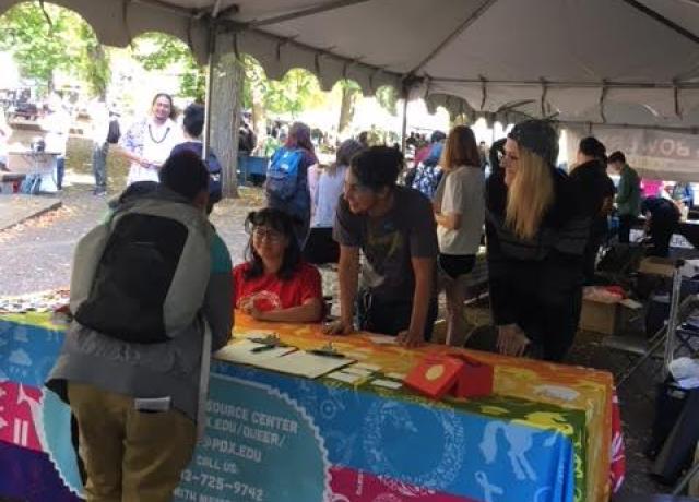 two students sit at a rainbow table with a QRC logo and lean in to chat with a table visitor