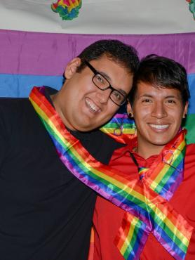 two brown queer people stand, heads together, with a rainbow sash around them, smiling in front of a rainbow flag.