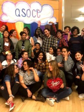 A group of BIPOC queer and trans people stand infront of a rainbow QSOCC sign, smiling and laughing