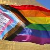 image of the progressive pride flag in the wind, a rainbow pride flag with a trangle added with black, brown, light blue, light pink, and white