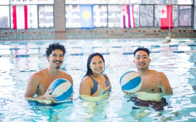 Three people standing in the Rec Center pool posing with beach balls.
