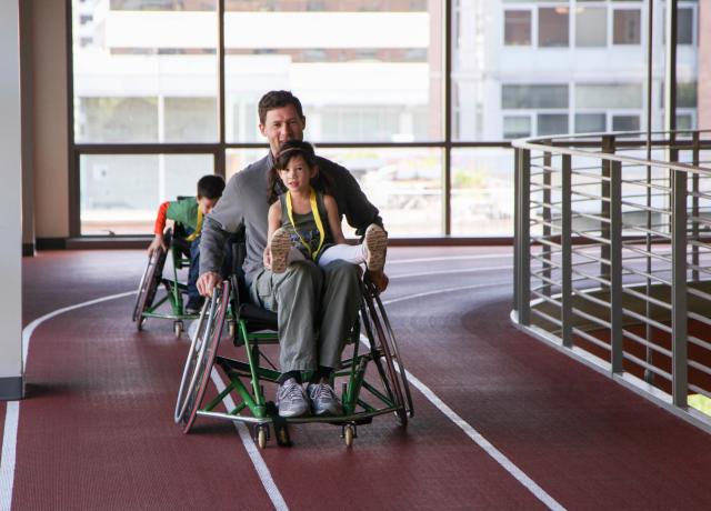 A parent rolls down the indoor track in sports chairs with two youth members.