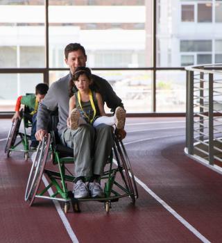 A parent rolls down the indoor track in sports chairs with two youth members.