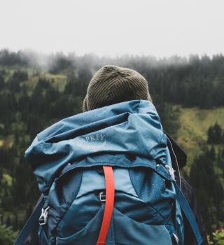 The image is of a person with a large blue backpack that has an orange strap, and the person is standing outside in a foggy forest.