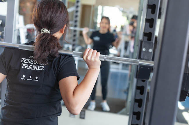 A personal trainer faces a mirror and lifts a barbell over her shoulders.