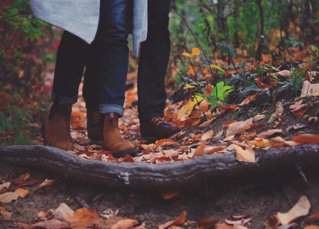 Two people in boots, walking on a forest trail with leaves and branches on the ground.
