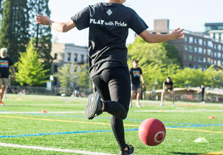 Person with a "Play with Pride" shirt is kicking a ball to teammates during the Pride Kickball Tournament in 2019 at Stott Field.