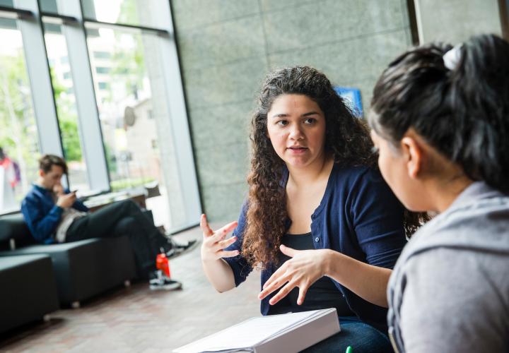 Two people discussing something in the ASRC lobby; one has a binder on her lap and her hands are raised in a gesture.