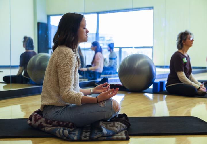 There is a person doing yoga in front of a large mirror, there is a grey yoga ball in the background. 