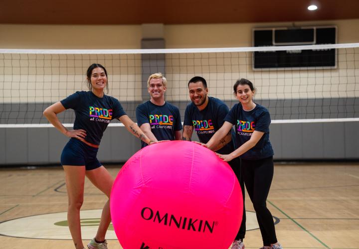 Players smile and pose next to giant pink volleyball wearing Pride shirts