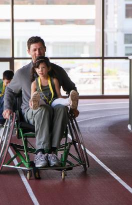 A youth member smiles and rides on the lap of a caregiver in a sports chair on the indoor track