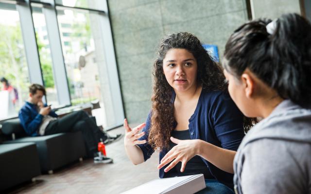 Two people discussing something in the ASRC lobby; one has a binder on her lap and her hands are raised in a gesture.