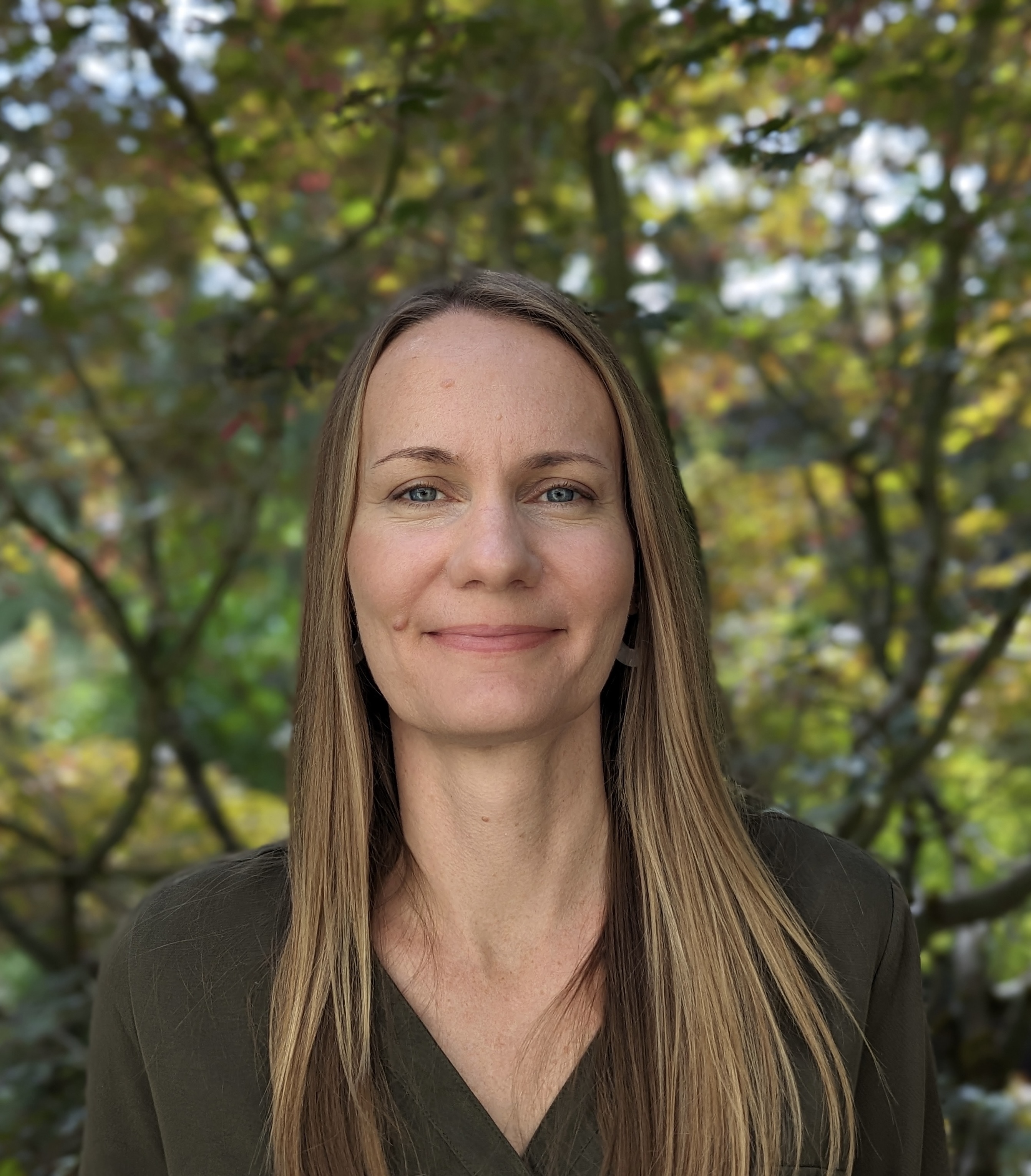 Woman with straight blonde hair faces camera smiling, with background of trees