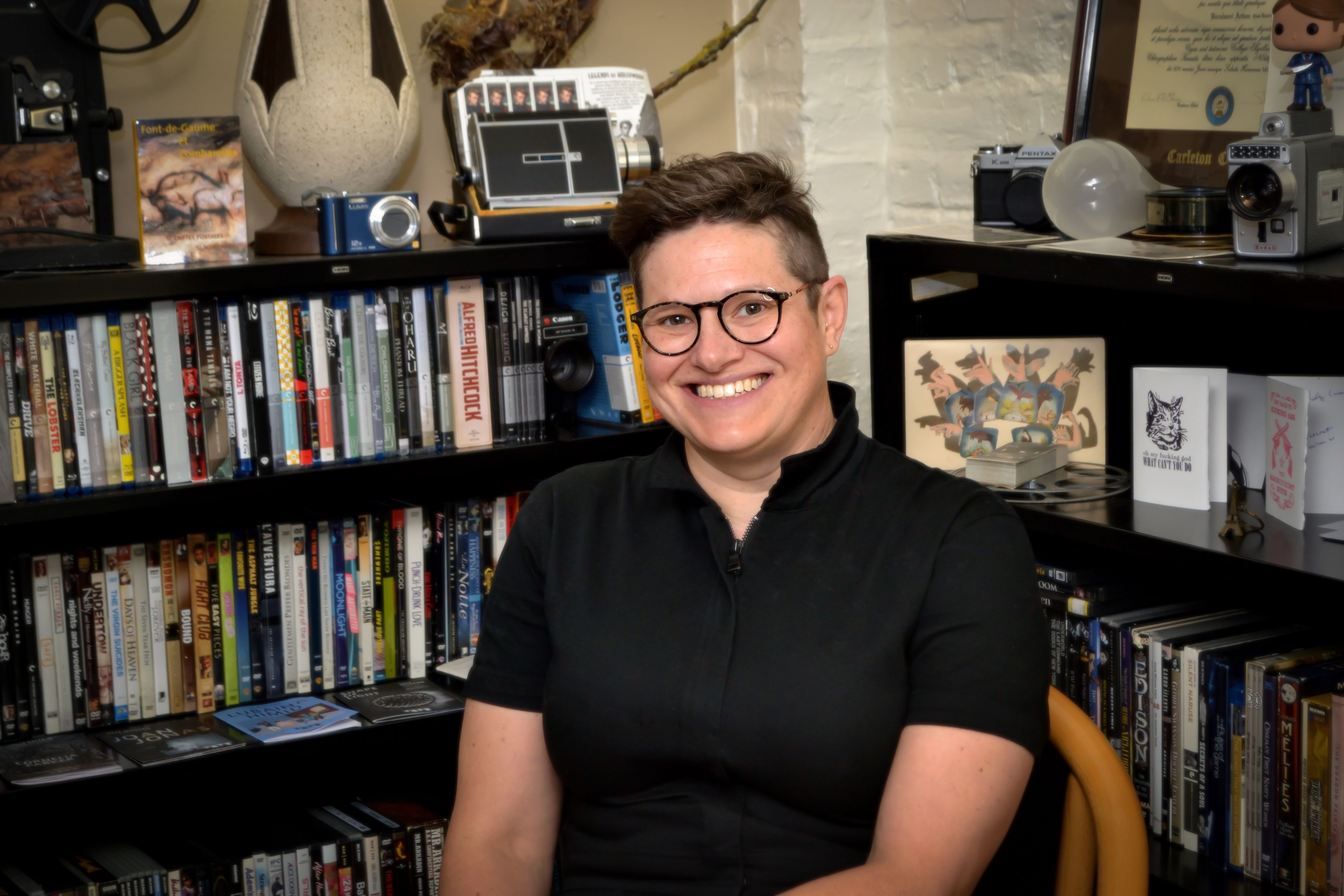Amy sits in a chair wearing a dark short-sleeved shirt, facing the camera and smiling, with books behind them