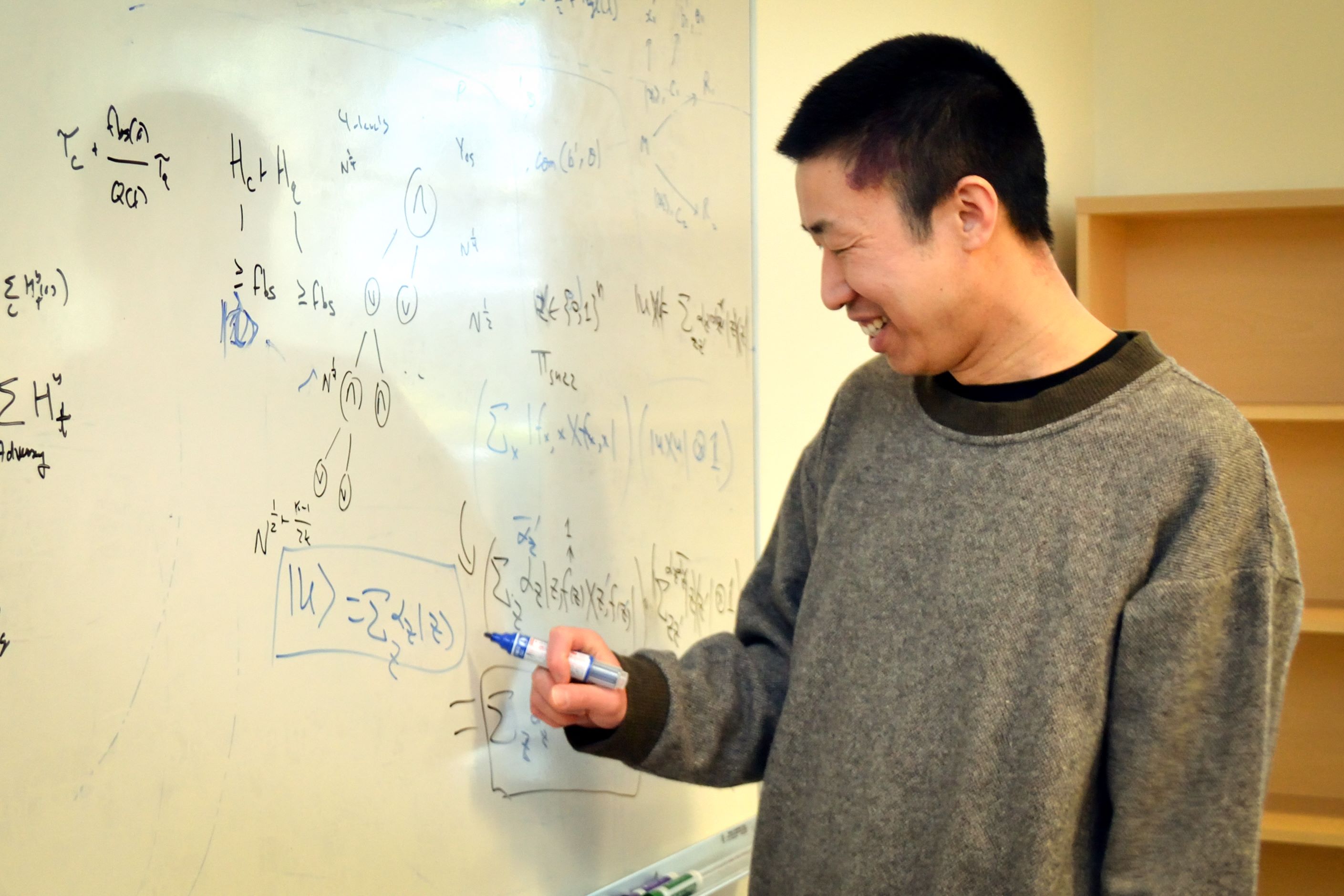 Fang Song faces a white board on which is written many calculations. He is smiling and wearing a gray sweater