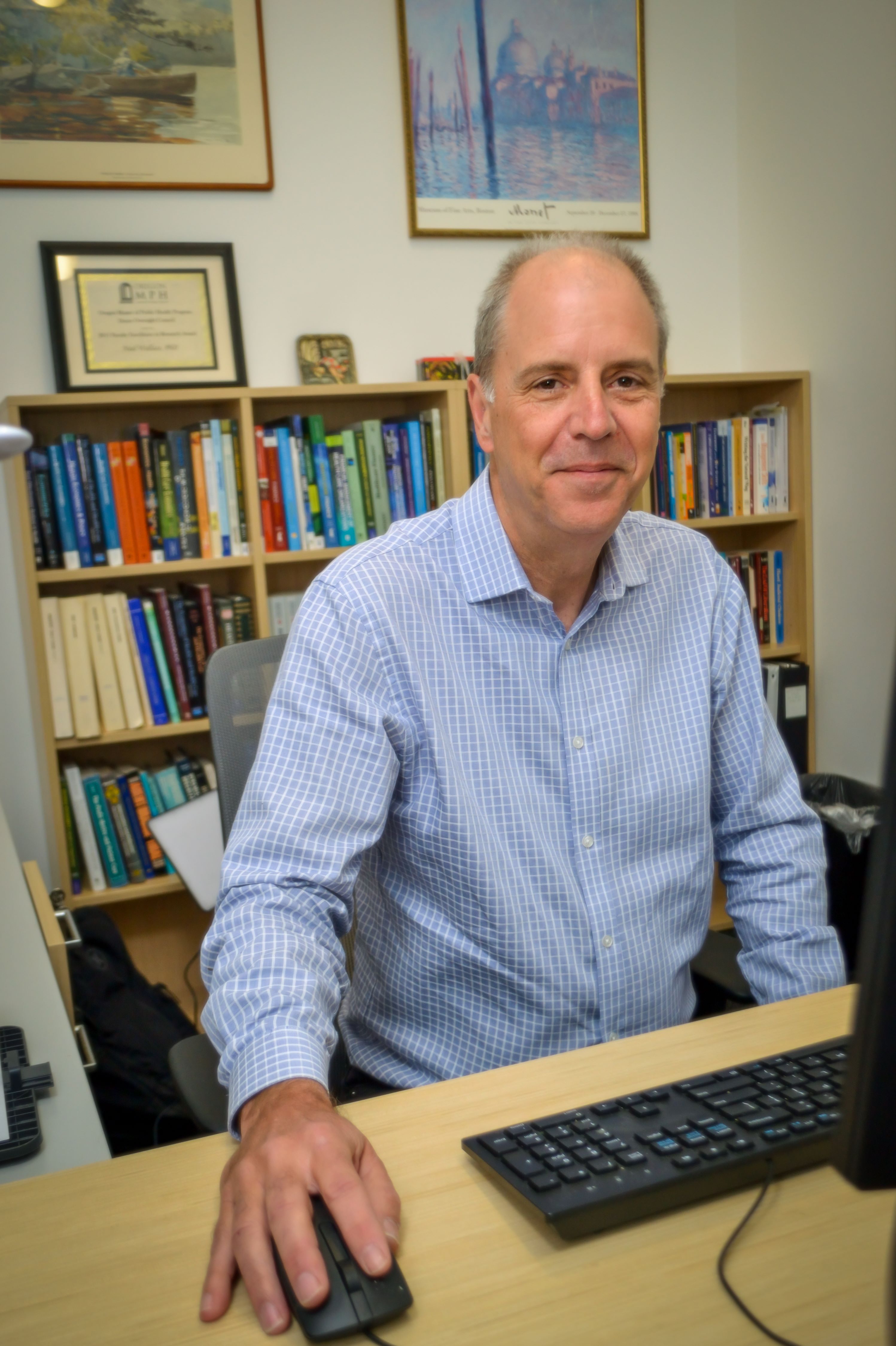 Neal sits at his desk, facing the camera and smiling. He wears a blue collared shirt and there are bookshelves behind him