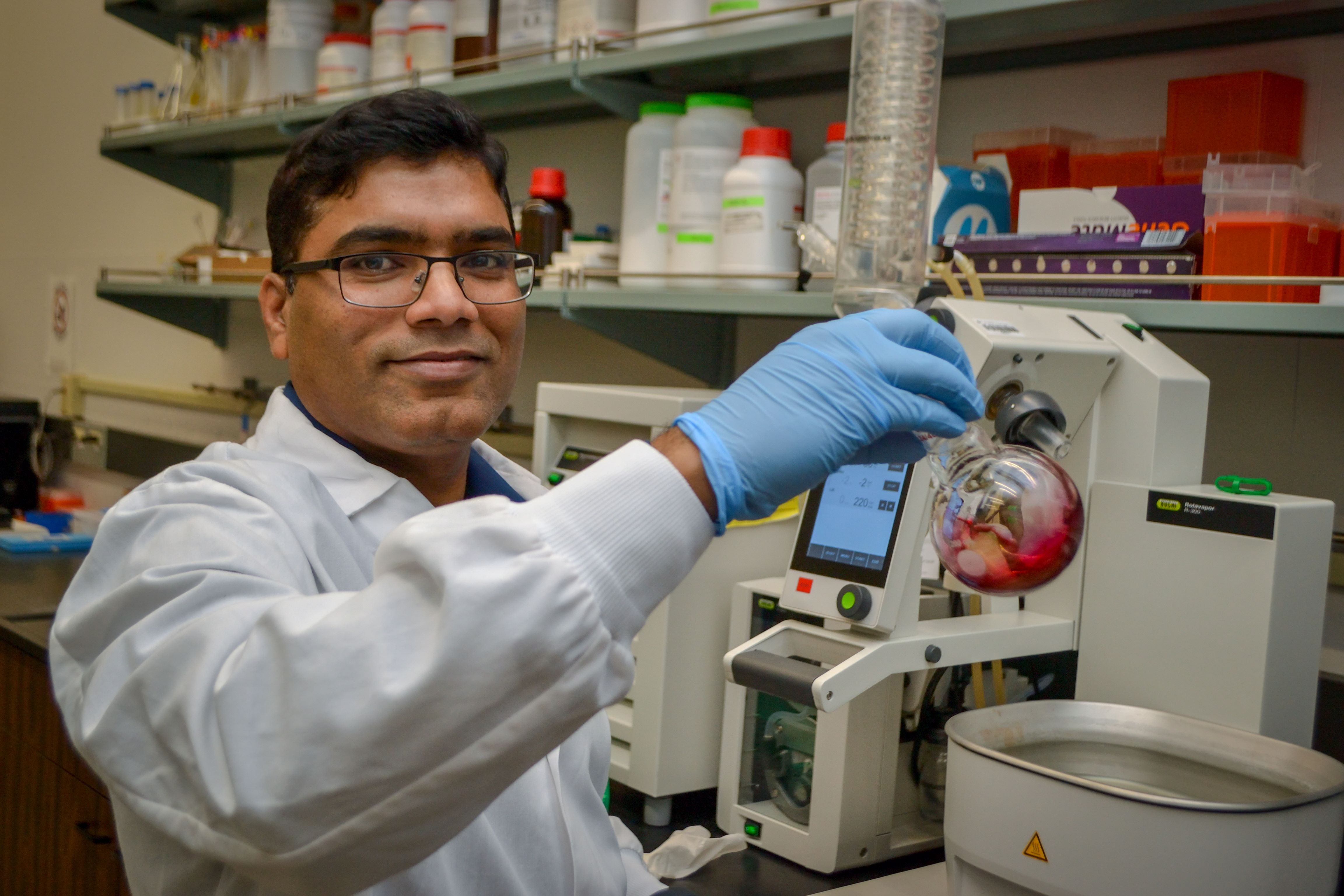 Papireddy Kancharla holds glass tube up, looking at camera. He wears a white lab jacket and is surrounded by lab equipment.