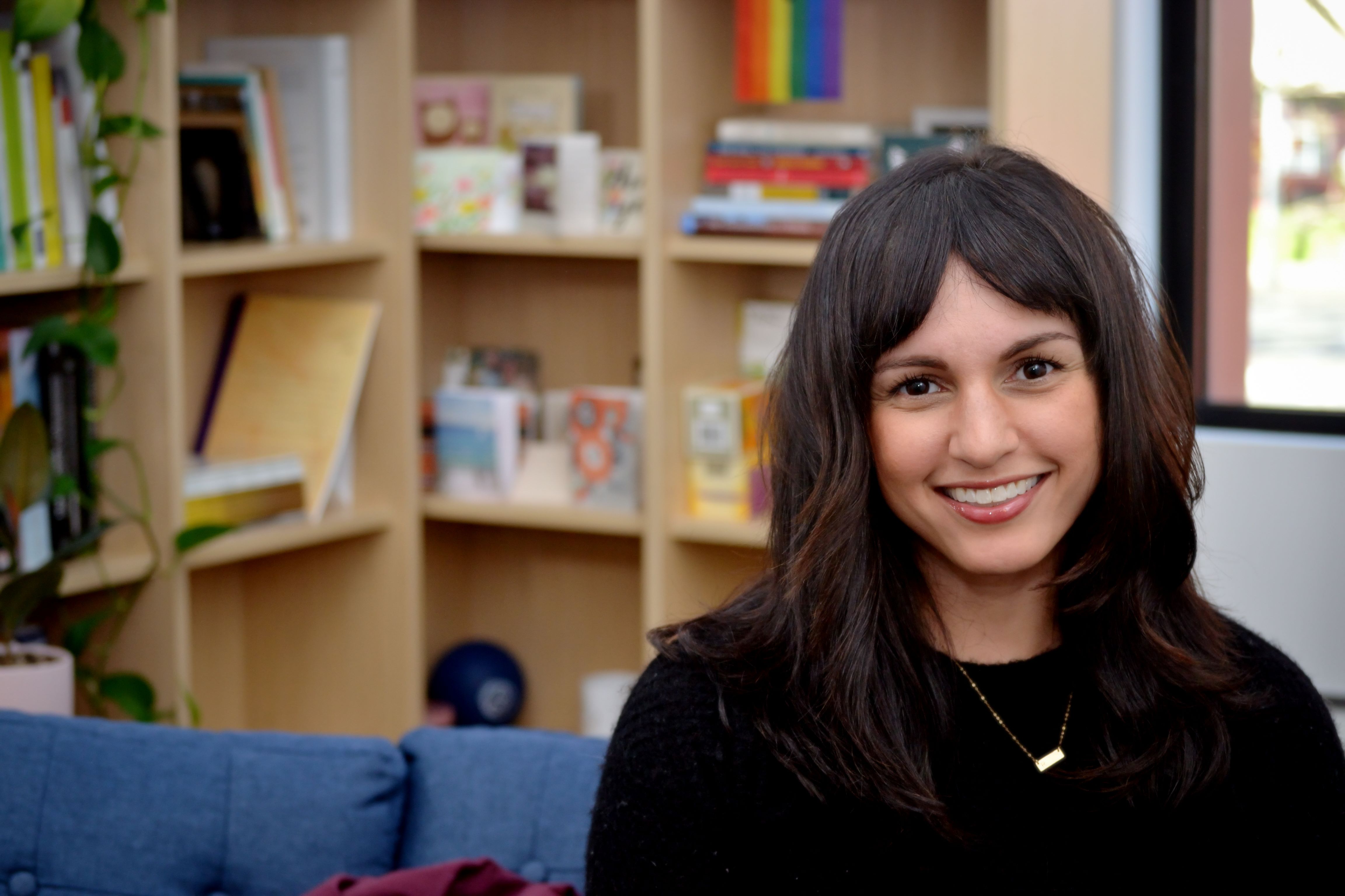 Rana sits to the right of the frame, facing the camera and smiling, with shelves of items behind her