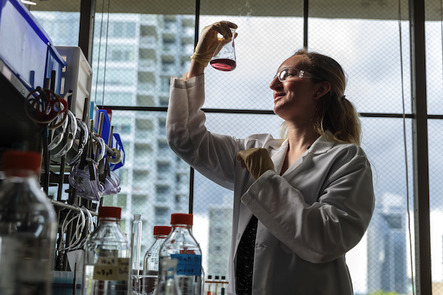 Light-skinned person with long blond hair, wearing a white lab coat and glasses, lifts a glass vial with colored liquid up to the light