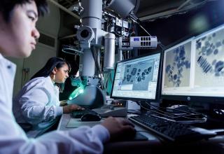 Two PSU researchers in labcoats looking at microscope and computer screens