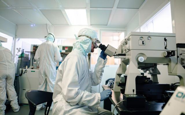 Students in cleanroom suits work in a high-tech laboratory. One person sits at a microscope examining samples, while others operate specialized equipment in the background, all wearing full protective gear including gloves, masks, and goggles.