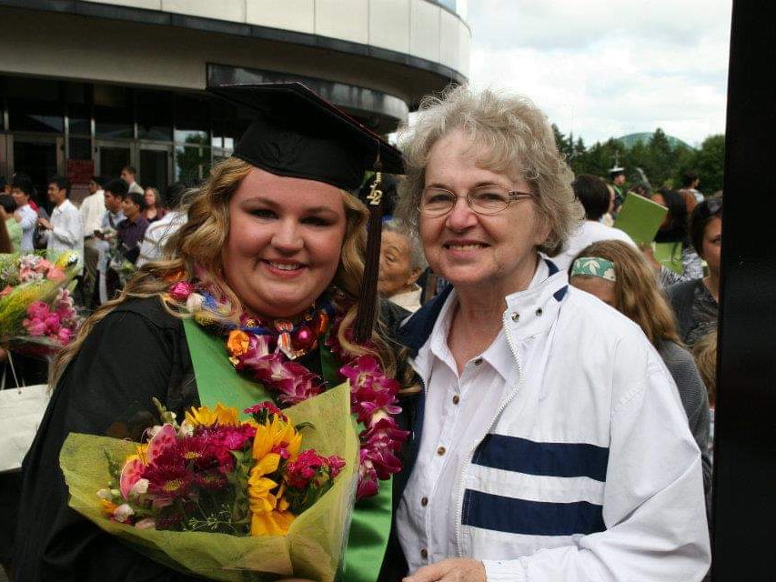 The image is of two people standing together, one of them is wearing graduation regalia. 