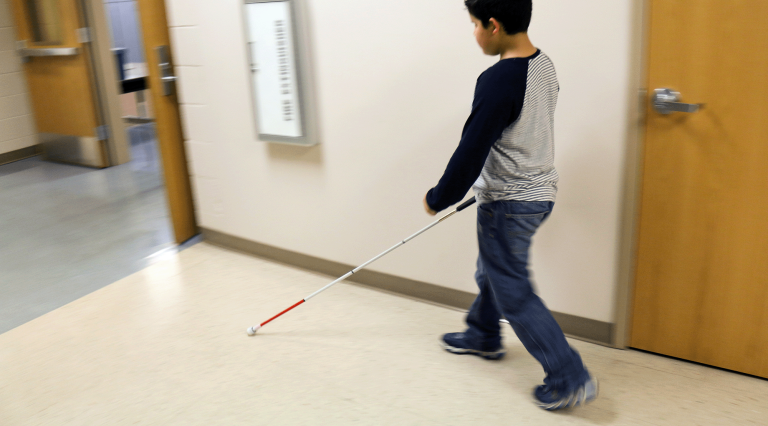 A young boy walks down a corridor using a white cane.