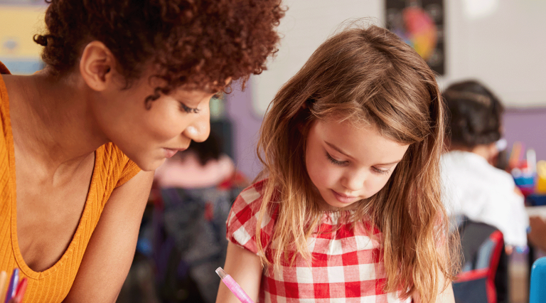 A smiling teacher with dark brown curls and an orange shirt assists a young child with her coursework.
