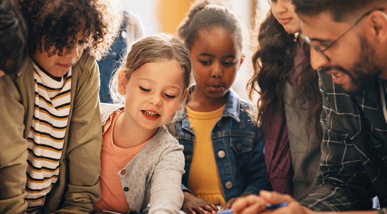 A teacher in a plaid shirt and glasses watches students complete an assignment.