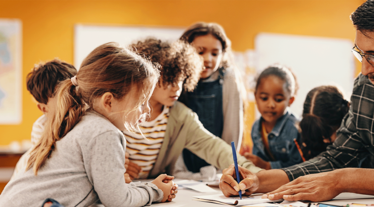 A male teacher in a grey plaid shrit and black glasses surrounded by a group of elementary school students.
