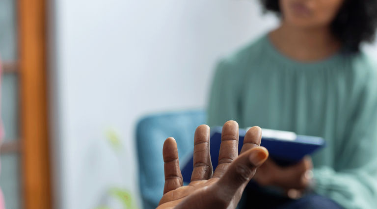 A counselor in a turquoise shirt holds a tablet and speaks with their patient, a man with dark skin and a pink shirt. Neither of their faces are visible.