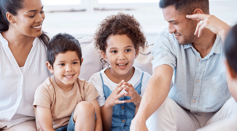 A mother, father, and two young children sit on a white couch together, smiling as they address a counselor.