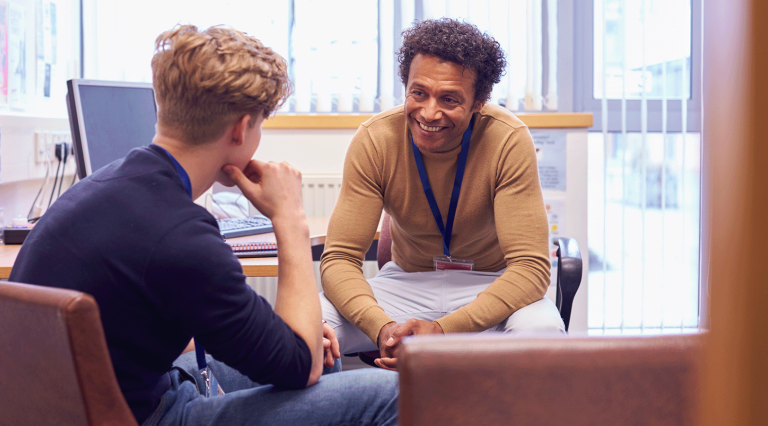 A school counselor with dark, curly hair and an ochre shirt smiles while advising a student with a blond undercut and dark blue clothes.
