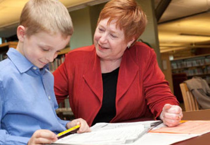The image is of Margaret Doherty helping a child with their homework at a table. 