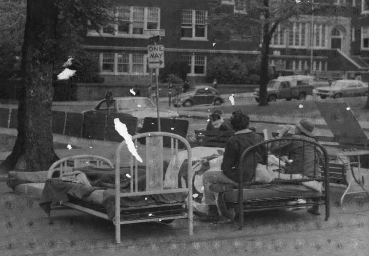 The image is of several beds in a parking lot, with a few people sitting on them.