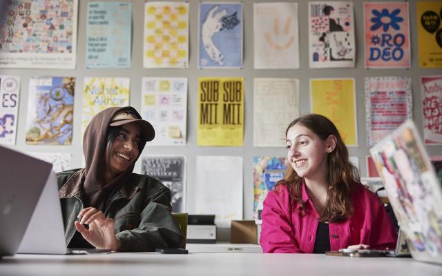 Two PSU students smile while working in a creative studio with posters behind them