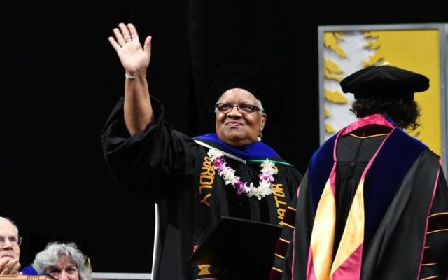 Avel Gordly wearing a graduation cap and gown waves to a crowd while wearing a flower lei