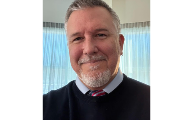 Dale Gaubatz smiling with short grey hair and a goatee wearing a blue shirt and striped tie
