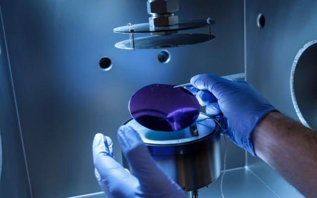 Gloved hands use tweezers to handle a circular semiconductor wafer inside a high-tech laboratory chamber