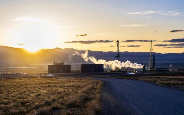 Geothermal research site with industrial equipment and steam rising against a sunset mountain backdrop