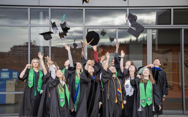 PSU graduates in black regalia and green stoles toss graduation caps into the air outdoors