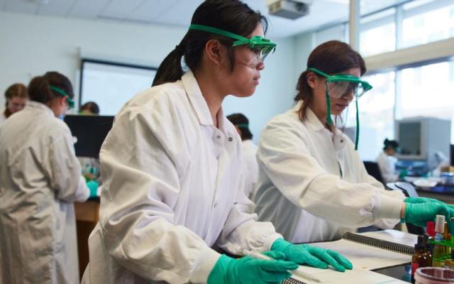 Two PSU researchers wearing lab coats and safety goggles work with equipment at a laboratory bench