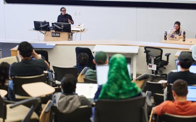 A PSU instructor speaking at a podium in a lecture hall filled with seated students using laptops