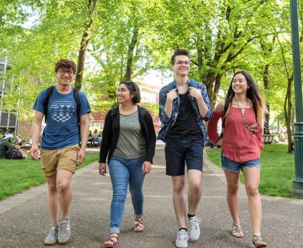 Four diverse students with backpacks walking and smiling together through the sunlit South Park Blocks on the Portland State campus