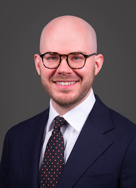 A professional headshot of a bald white man with acetate glasses and short beard in a white shirt, tie, and dark navy business suit.