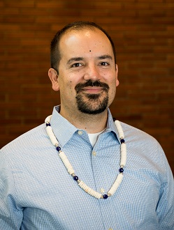 Headshot of Native American man in a blue shirt in front of a brick wall