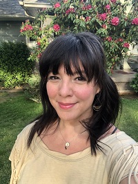 White woman with black hair and a yellow blouse smiling in front of a background of flowers