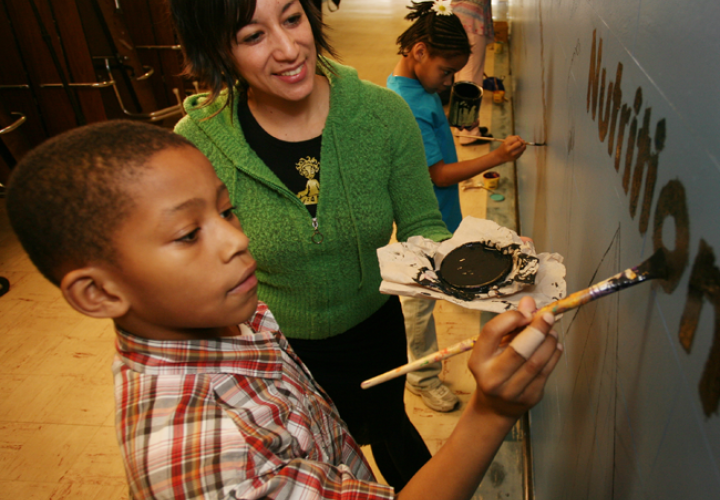 Photo of a person helping a child paint the word "Nutrition" on a wall.