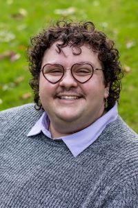 Professional photo of grad student Madi Lou. They are wearing a grey sweater and heartshaped glasses.