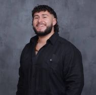 Photo of a graduate student. They are wearing a black button down shirt and is sporting a mullet and a beard